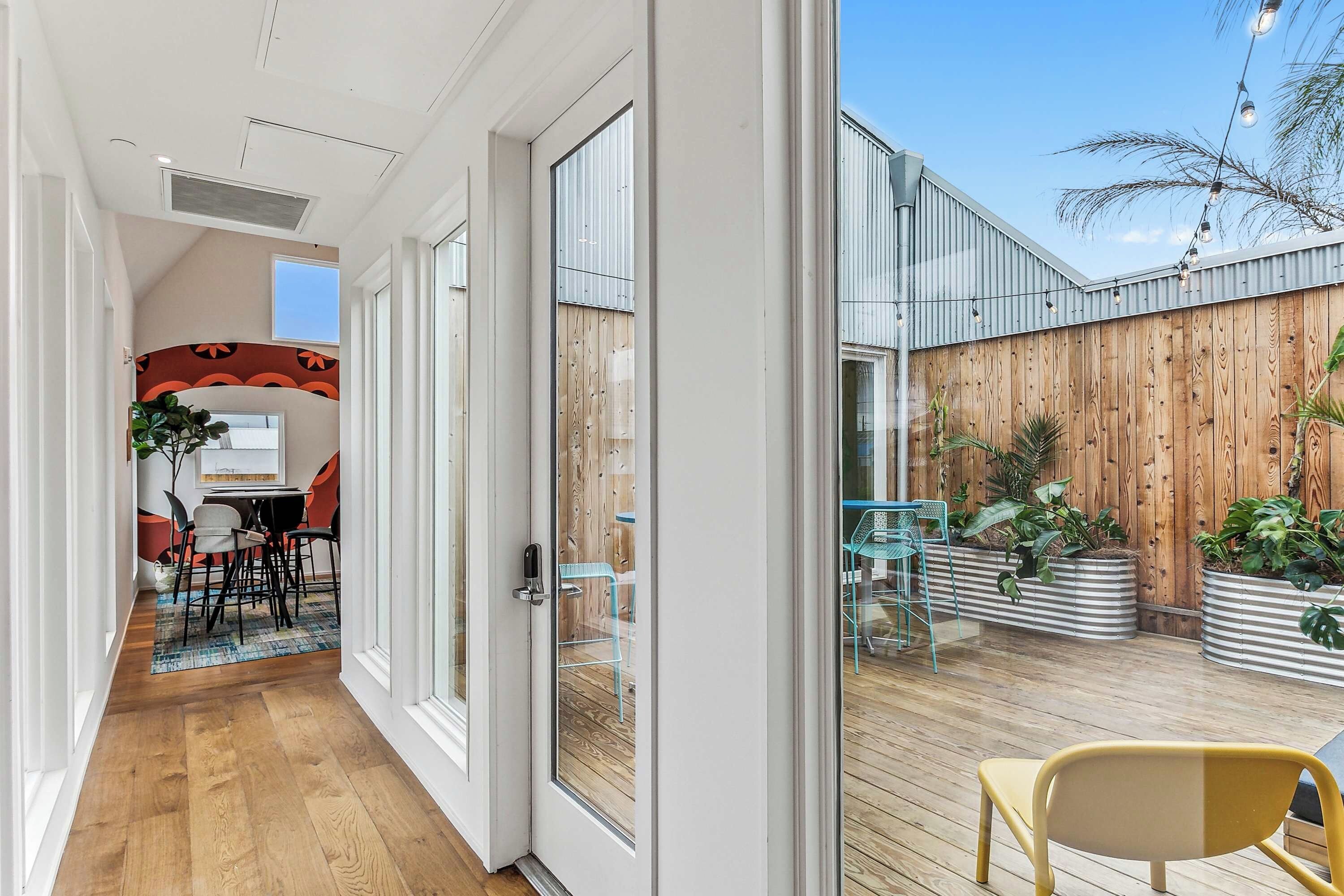 A modern interior with a wooden floor, a dining area with a table and chairs, and a view of a wooden fence and plants outside.