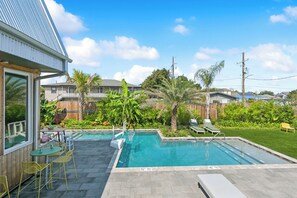 A backyard with a swimming pool, a patio with chairs, and a wooden fence.