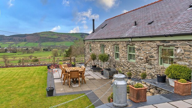 A stone house with a tiled roof, a patio with a table and chairs, and a view of the countryside. - Moelis Barn (Llandrillo)