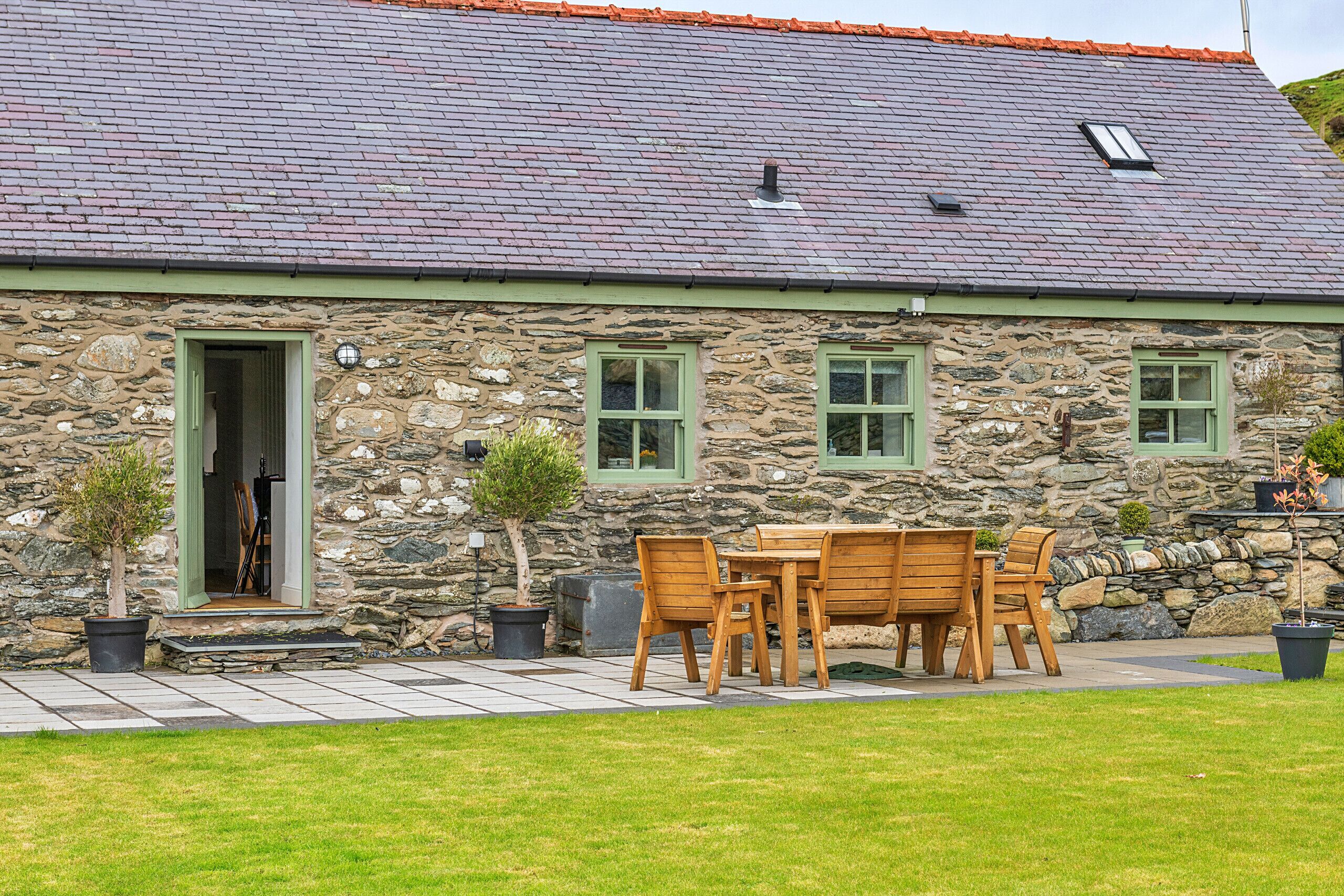 A stone building with a tiled roof, a wooden table and chairs outside, and a small tree in a pot.