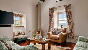 A cozy living room with a fireplace, green sofas, a wooden coffee table, and floral curtains. - Moelis Barn (Llandrillo)