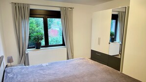 Ein Schlafzimmer mit einem Bett, einem Fenster mit Vorhängen, einem Heizkörper, einer Zimmerpflanze und einem Spiegel.