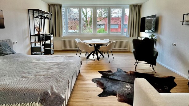 Ein modernes Schlafzimmer mit einem großen Bett, einem Esstisch mit Stühlen, einem Fernseher und Blick ins Freie.