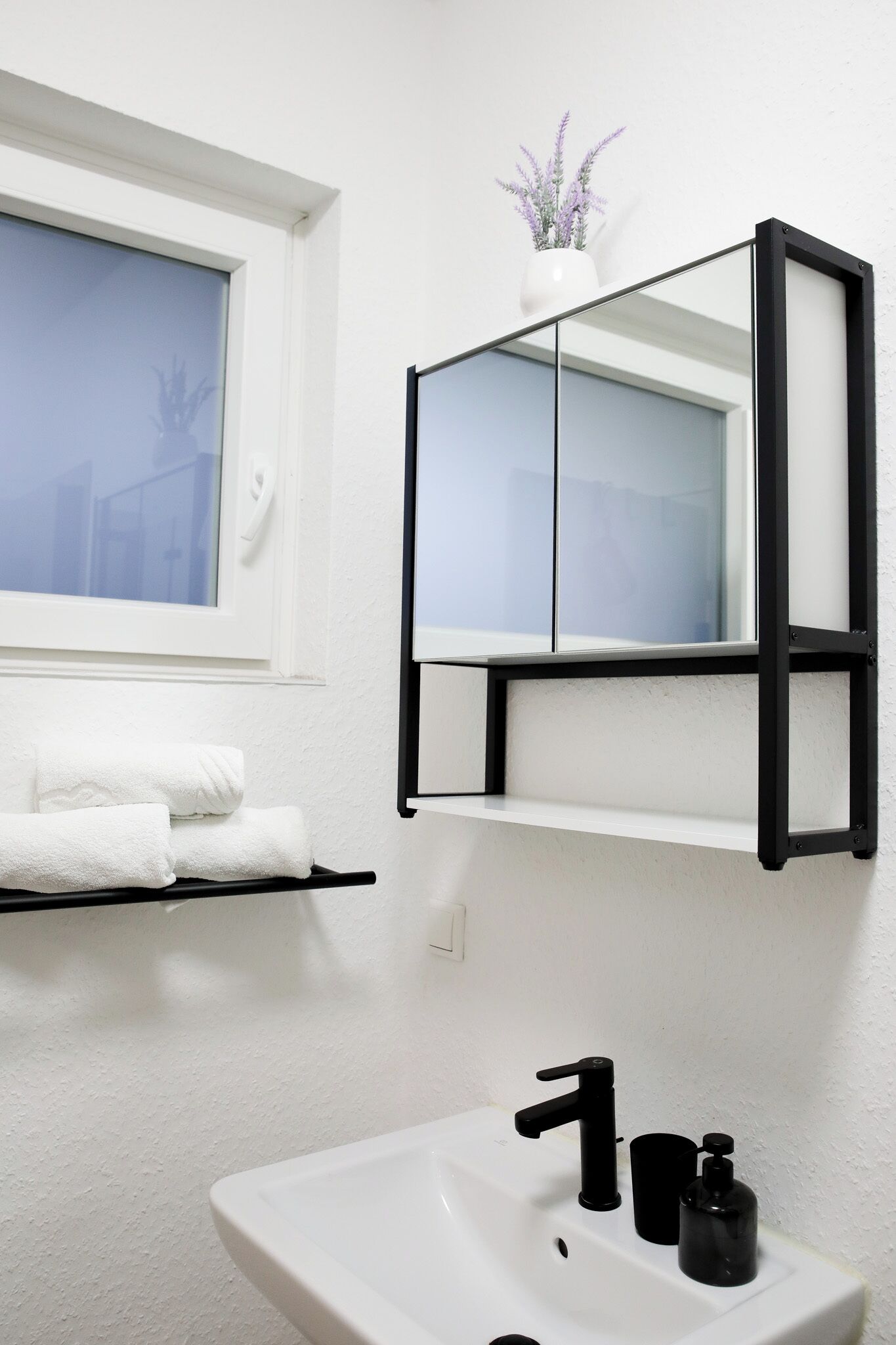 A bathroom with a white sink, black faucet, and a mirror with a black frame. There is a window with a view of a building outside.