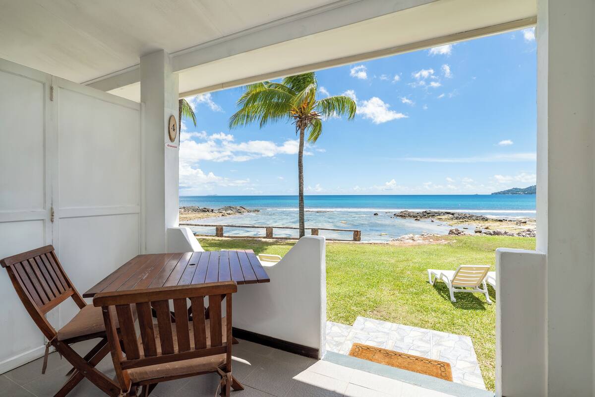 A covered patio with a wooden table and chairs, a palm tree, and a view of the ocean.