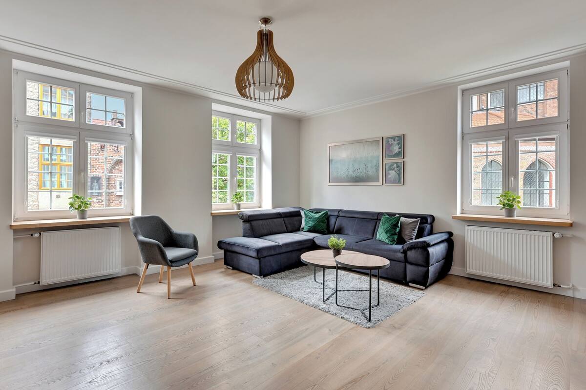 Ein modernes Wohnzimmer mit einer Eckcouch, einem runden Couchtisch und einem Sessel. Große Fenster bieten Blick auf umliegende Gebäude, unter jedem Fenster befindet sich eine Heizung.