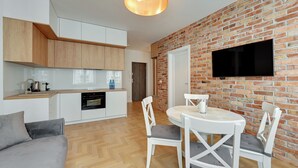 Une cuisine moderne avec des meubles blancs, un coin repas doté d’une table et de chaises blanches, et un mur en briques sur lequel est fixée une télévision.