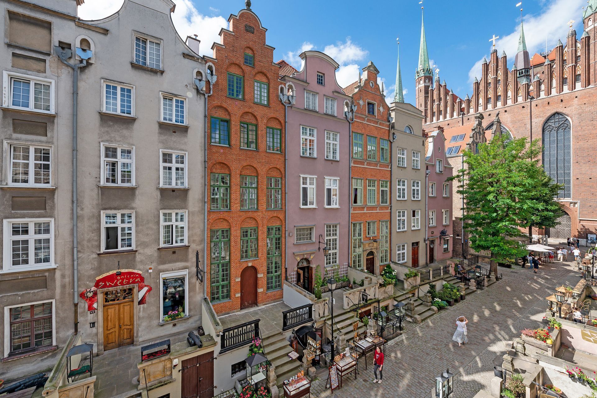 A historic European street with multi-story buildings, a church with a prominent spire, and a cobblestone plaza with outdoor seating.