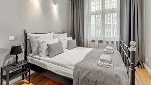 Un lit bien fait, avec du linge de lit blanc et des oreillers gris, une table de chevet avec une lampe et une fenêtre ornée de rideaux légers.