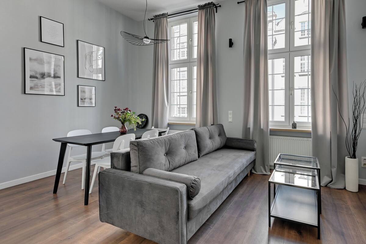 Ein modernes Wohnzimmer mit grauem Sofa, einem Glastisch und einem Essbereich mit weißem Tisch und Stühlen.