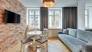 Un salon moderne avec un mur en briques, un téléviseur à écran plat, une table de salle à manger ronde avec des chaises, un canapé gris et de grandes fenêtres avec des rideaux.