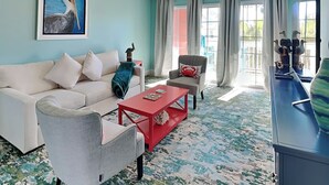 A living room with a white sofa, grey armchair, and red coffee table. There is a painting of a bird on the wall, a ceiling fan, and large windows with curtains.