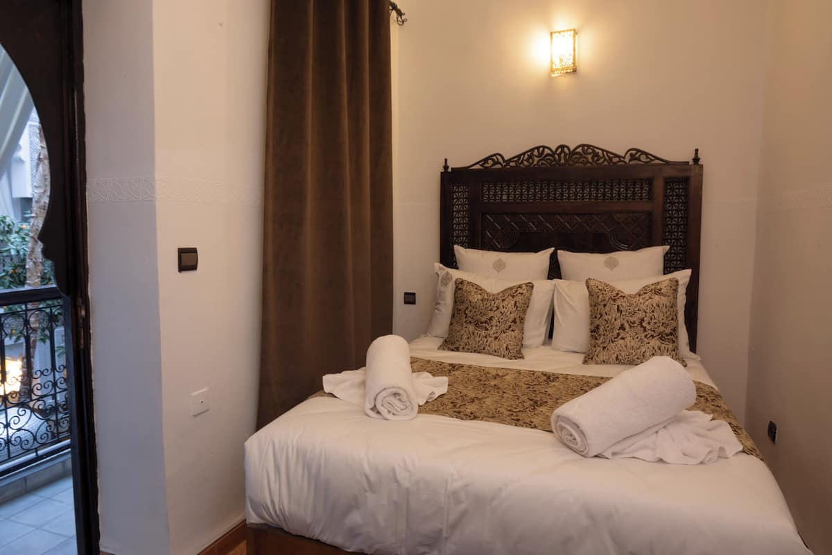 A neatly made bed with white linens and rolled towels, a patterned headboard, and a balcony with a wrought iron railing.
