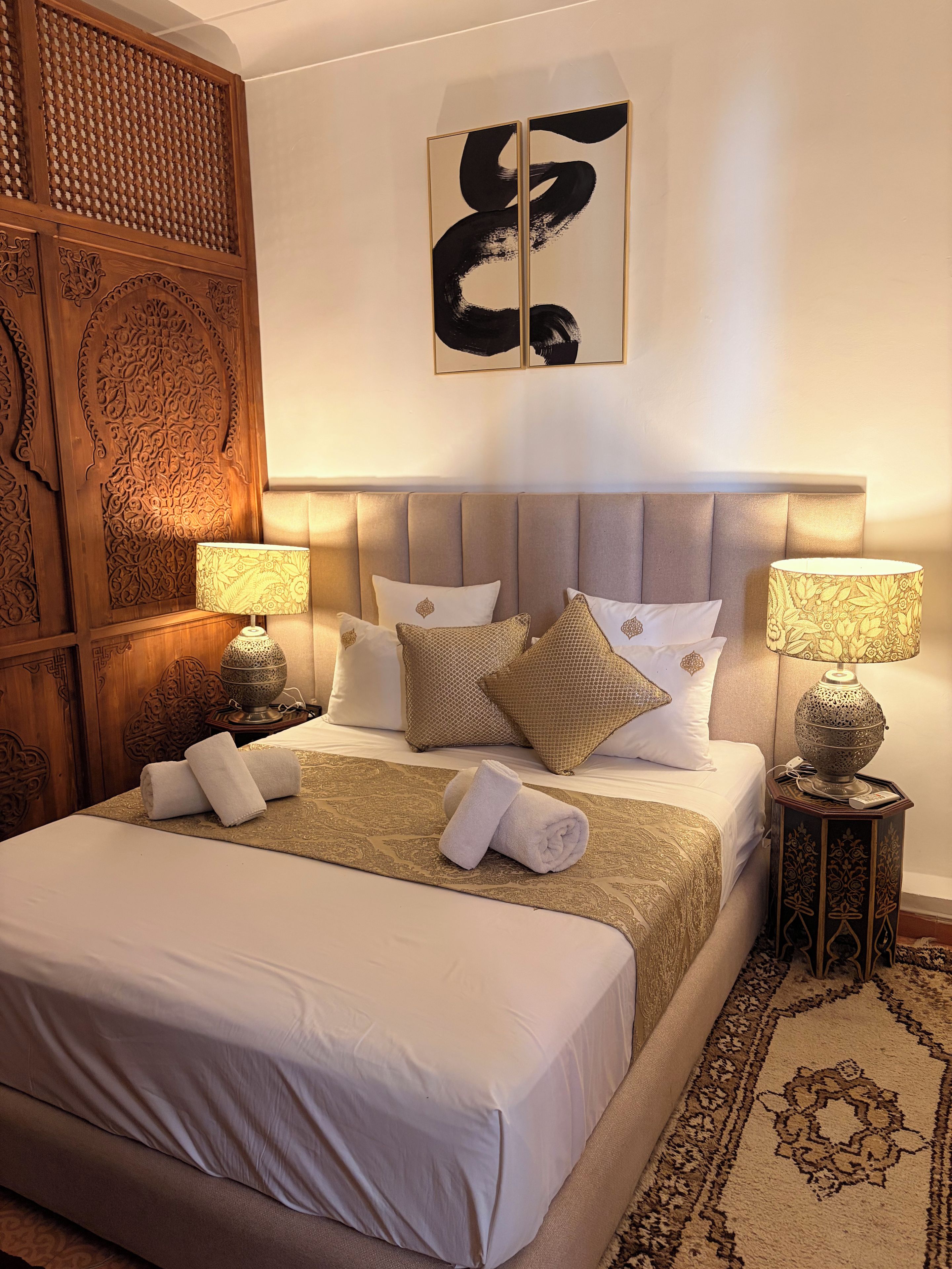 A well-appointed hotel room with a large bed, multiple pillows, a decorative headboard, two bedside lamps, and a patterned rug.