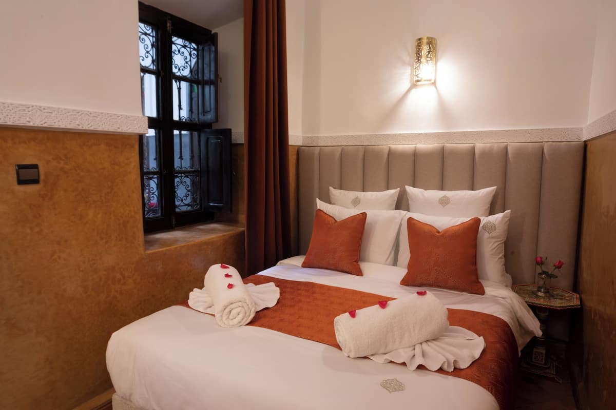 A hotel room with a large bed, a headboard, a bedside table with a vase of flowers, and a window with curtains.
