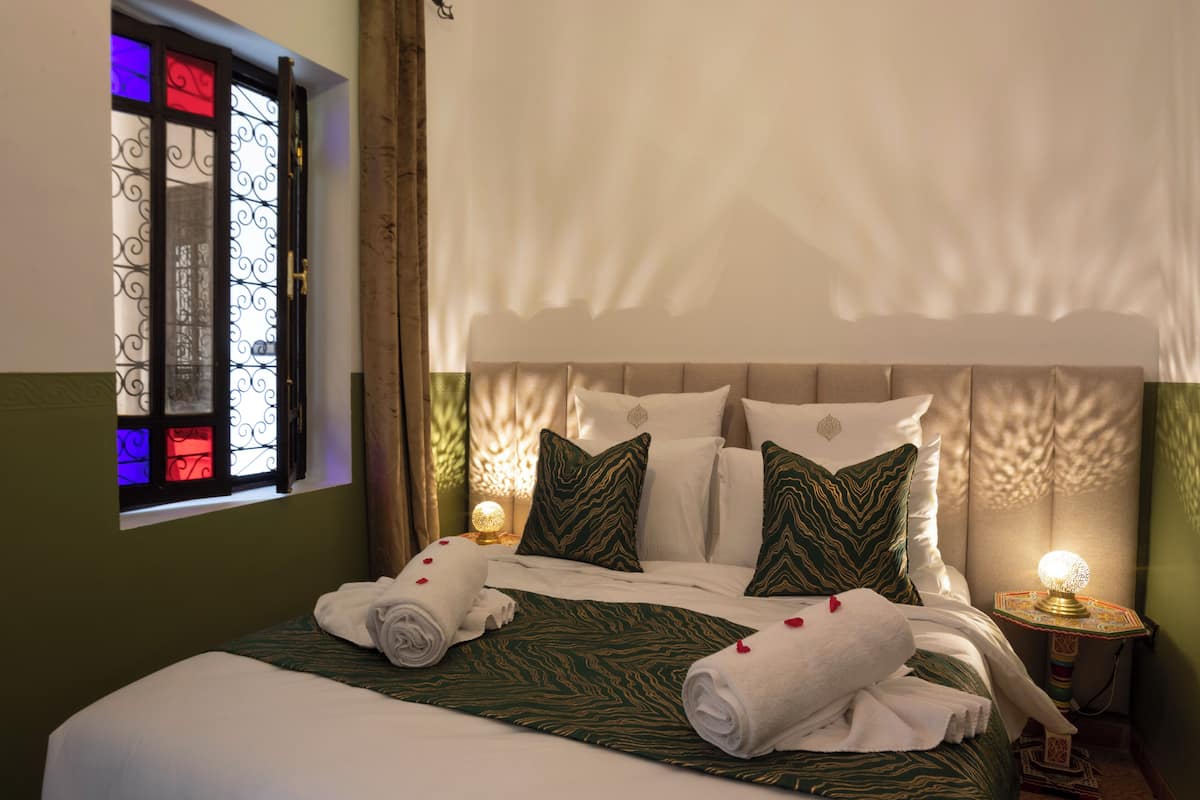 A neatly made bed with green and white bedding, a window with decorative iron bars, and a bedside lamp.