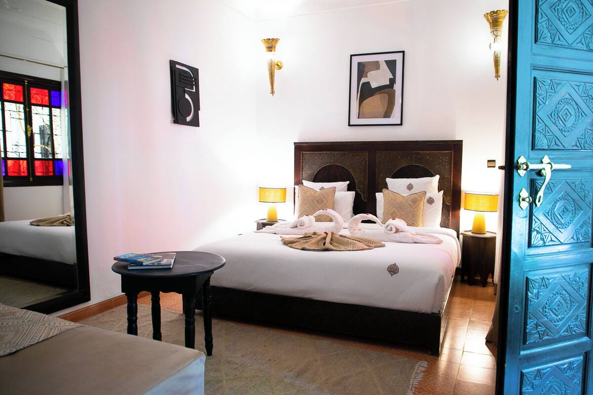 A hotel room with a bed, bedside tables, a small table, a mirror, and a blue door.