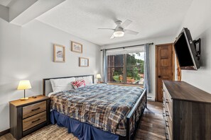 Une chambre à coucher comprenant un lit, des tables de chevet, un ventilateur de plafond, un téléviseur fixé au mur et une fenêtre avec des rideaux.