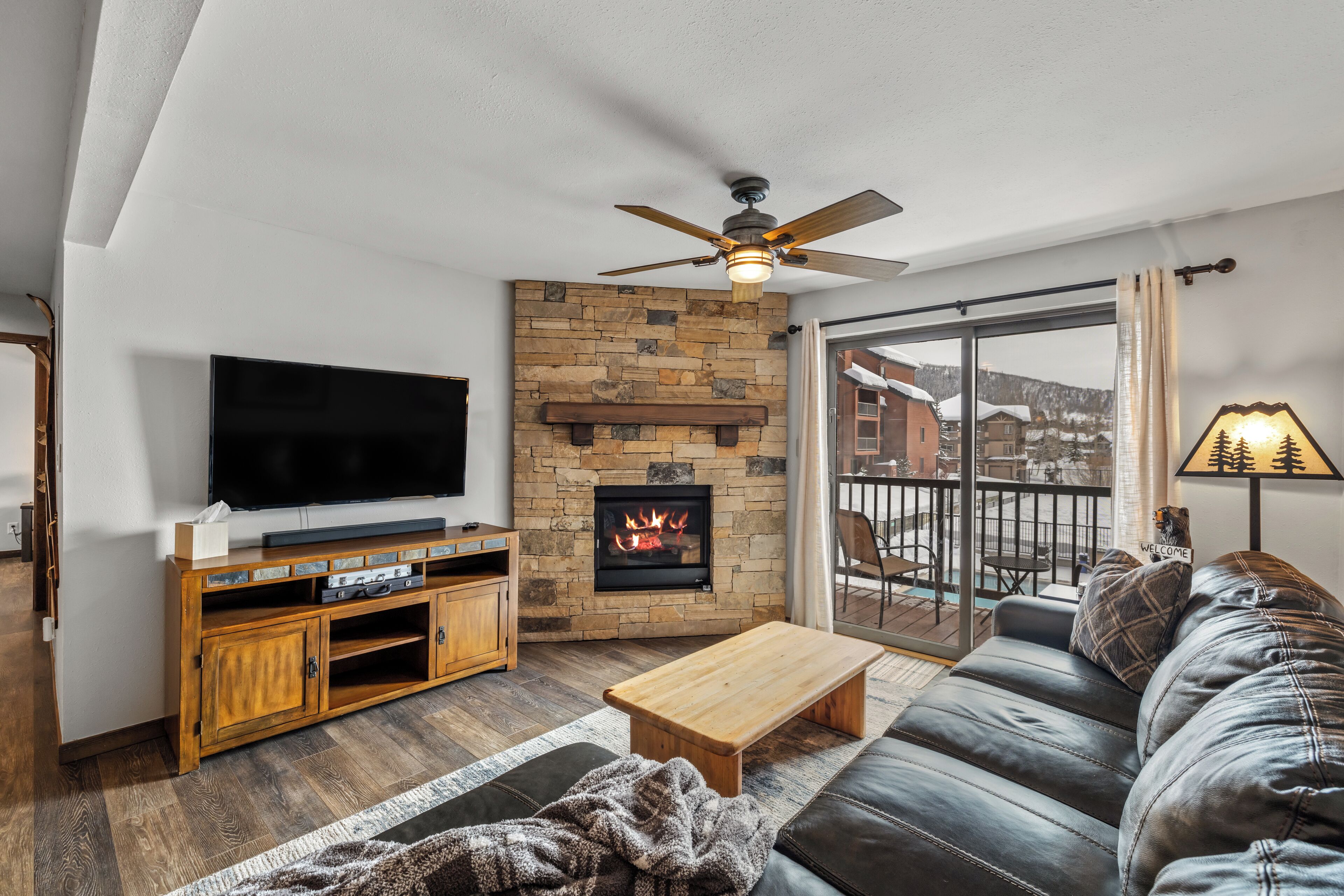 Ein Wohnzimmer mit Kamin aus Stein, einem Flachbildfernseher, einer Holz-Unterhaltungseinheit, einem Ledersofa und einem Balkon mit Blick auf schneebedeckte Häuser.