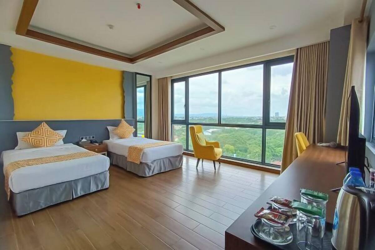 A hotel room with two beds, a large window with a view, a desk with amenities, and a yellow chair.