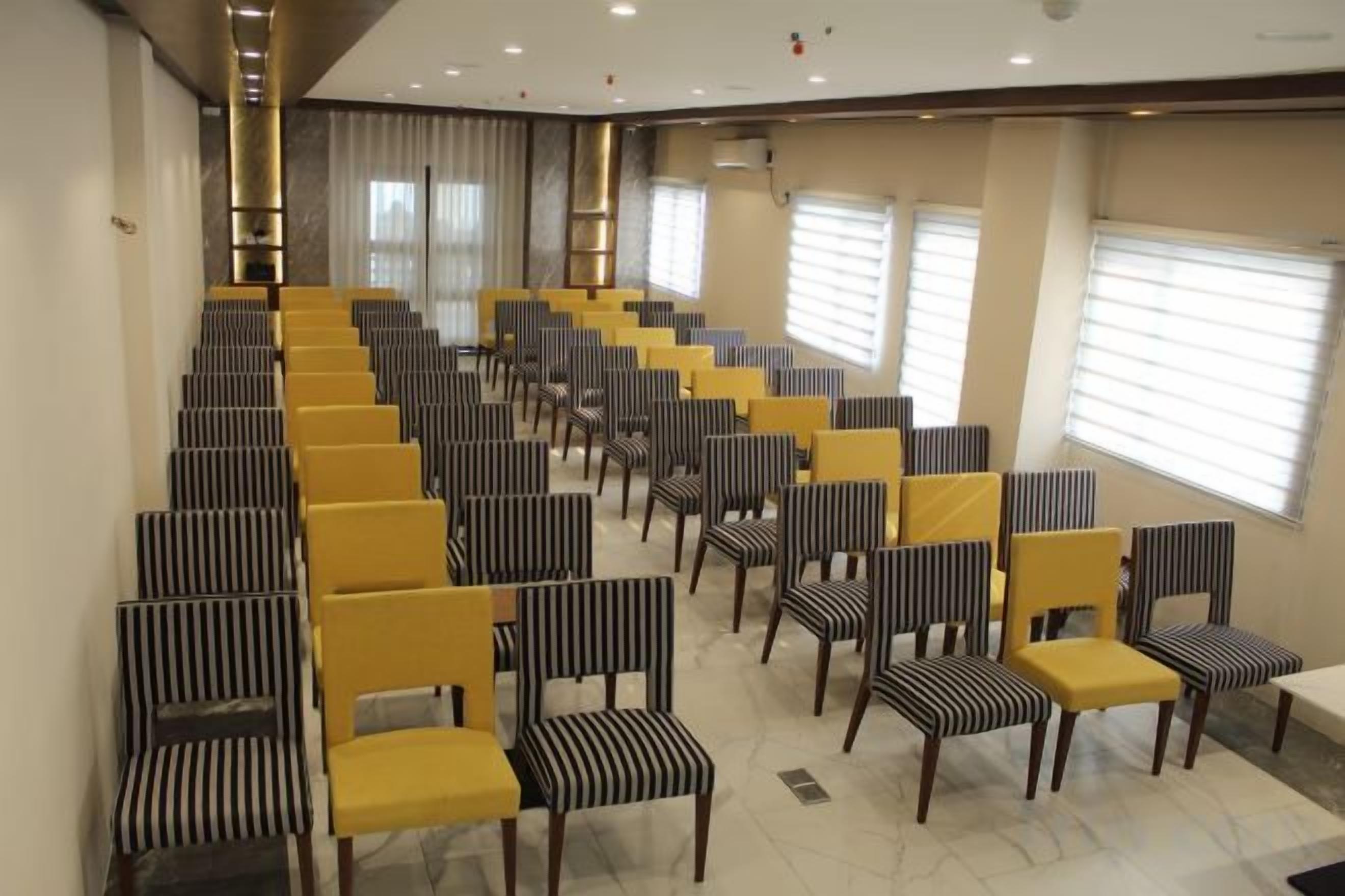 A conference room with rows of chairs and tables, large windows with blinds, and a high ceiling with recessed lighting.