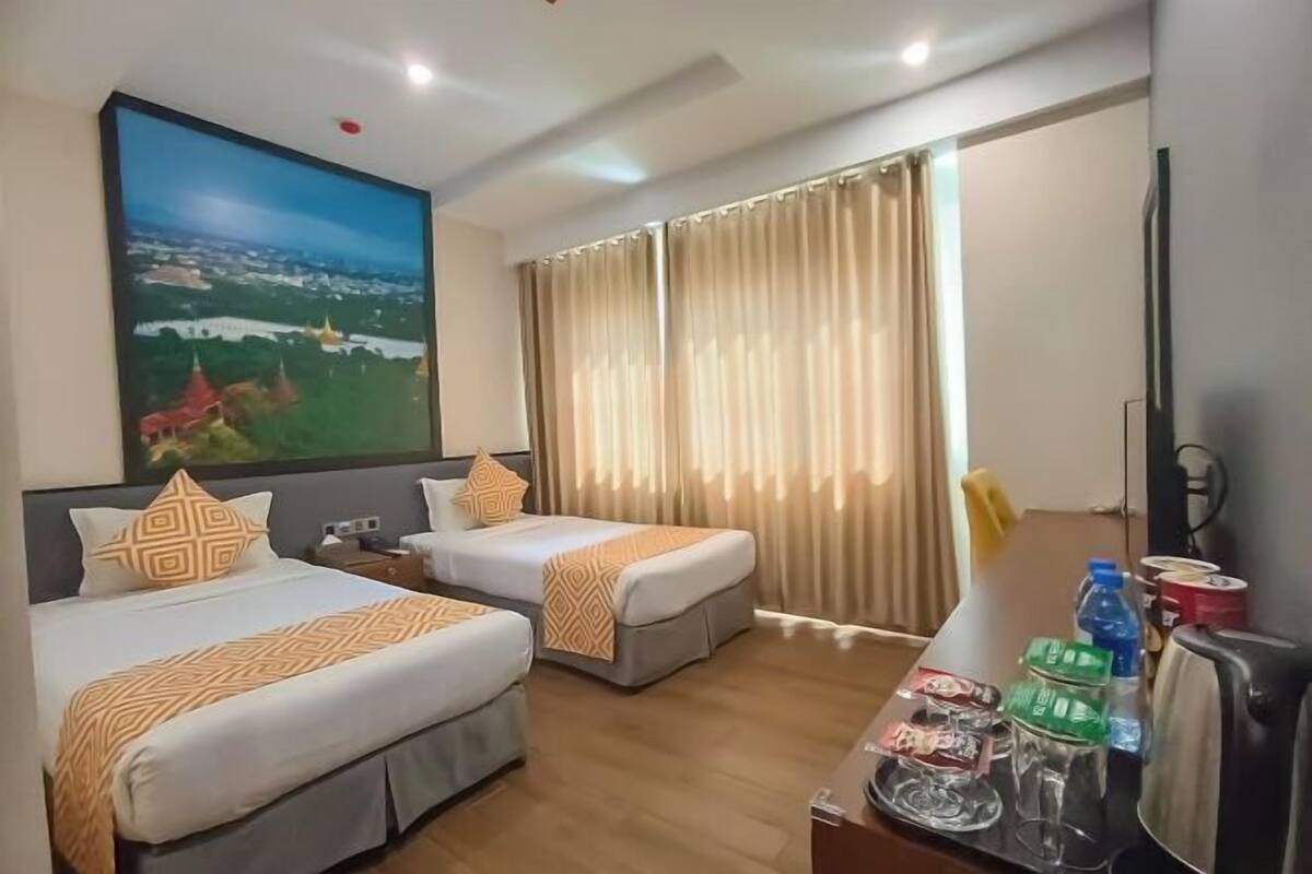 A hotel room with two beds, a large painting, a desk with a kettle, and curtains.