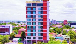 A multi-story hotel with a red and white facade, multiple windows, and a view of surrounding buildings and greenery. - Holiday Pier Hotel (Mandalay)