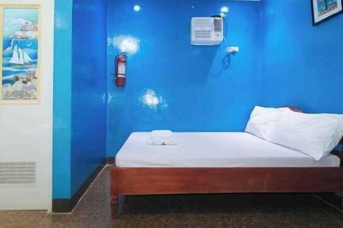 Une petite pièce comprenant un lit simple, un extincteur, un appareil de climatisation et un tableau encadré accroché au mur.