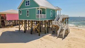 A green wooden house with a balcony on stilts, a staircase, and a view of the ocean. - New Construction! Beachfront, 3 Bedroom/3.5 Bath, Pool, Hot Tub, Pet Friendly (Dauphin Island)