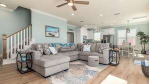 A modern living room with a sectional sofa, a coffee table, and a rug. There is a staircase with a wooden railing and a ceiling fan. - New Construction! Beachfront, 3 Bedroom/3.5 Bath, Pool, Hot Tub, Pet Friendly (Dauphin Island)