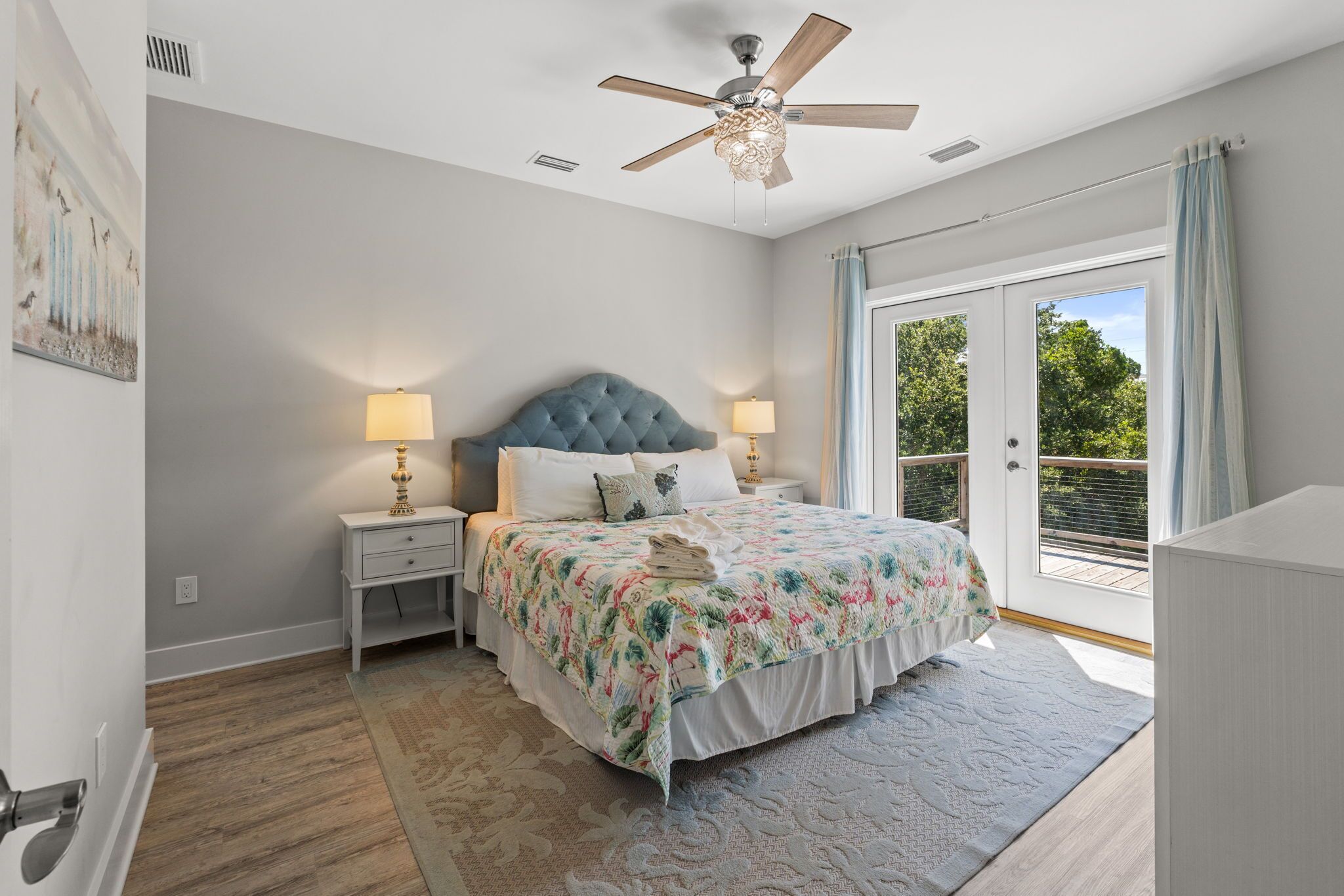 A bedroom with a large bed, two bedside tables with lamps, a ceiling fan, and a view of the outdoors through a sliding glass door.