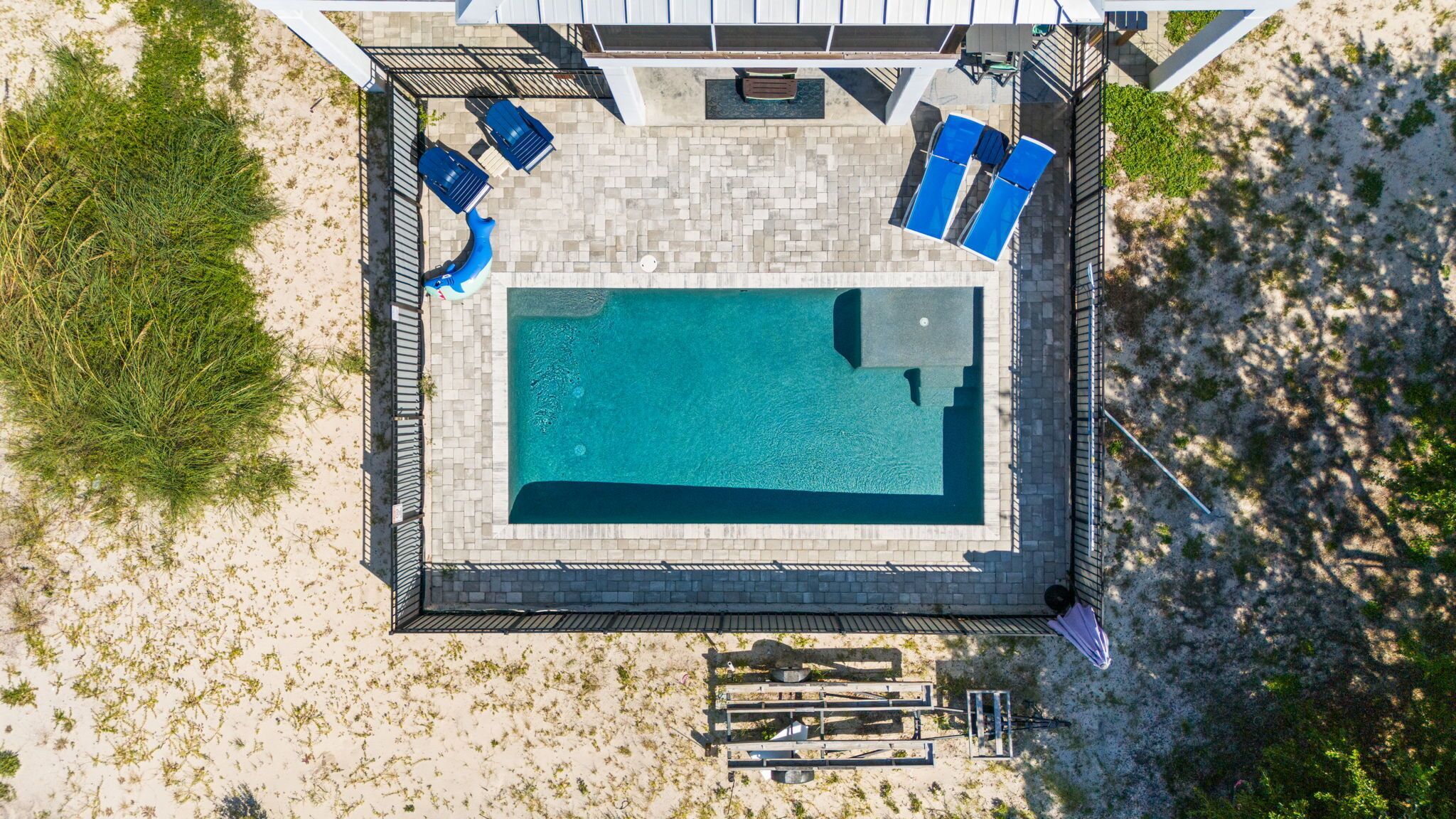 An aerial view of a rectangular swimming pool with a tiled deck, surrounded by a metal fence. There are lounge chairs and a barbecue grill visible.