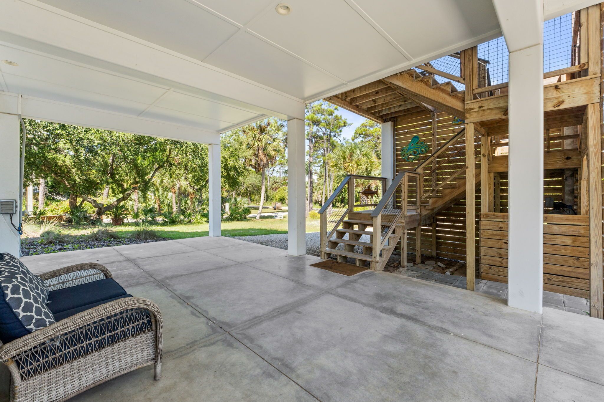 A covered patio area with a wicker sofa, a wooden staircase, and a view of a lush garden.