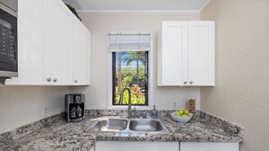 A kitchen with a granite countertop, stainless steel sink, white cabinets, a window with a view of greenery, and a coffee maker. - Kona Bali Kai#164:Stunning Remodeled Studio with Ocean Views: Walk to Beach! (Kailua Kona)