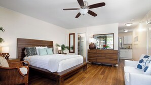 A bedroom with a wooden bed, a ceiling fan, a nightstand, a wicker chair, a white sofa, and a wooden dresser. - Kona Bali Kai#164:Stunning Remodeled Studio with Ocean Views: Walk to Beach! (Kailua Kona)