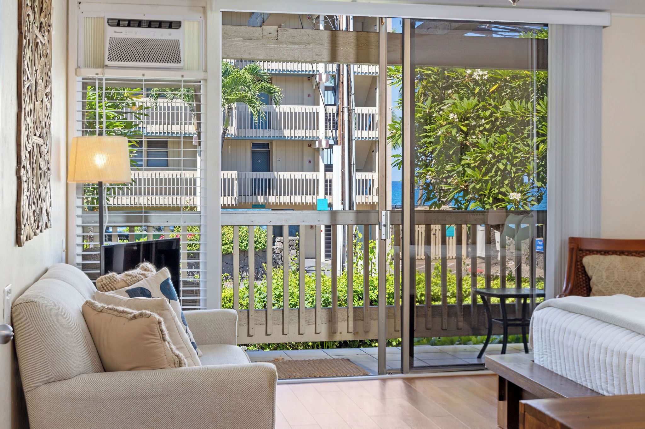 Ein Hotelzimmer mit einer Couch, einem Bett, einem Tisch und Blick auf einen Balkon mit Pflanzen sowie ein Gebäude im Hintergrund.