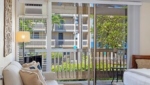 A hotel room with a sofa, a bed, a table, and a view of a balcony with plants and a building in the background. - Kona Bali Kai#164:Stunning Remodeled Studio with Ocean Views: Walk to Beach! (Kailua Kona)