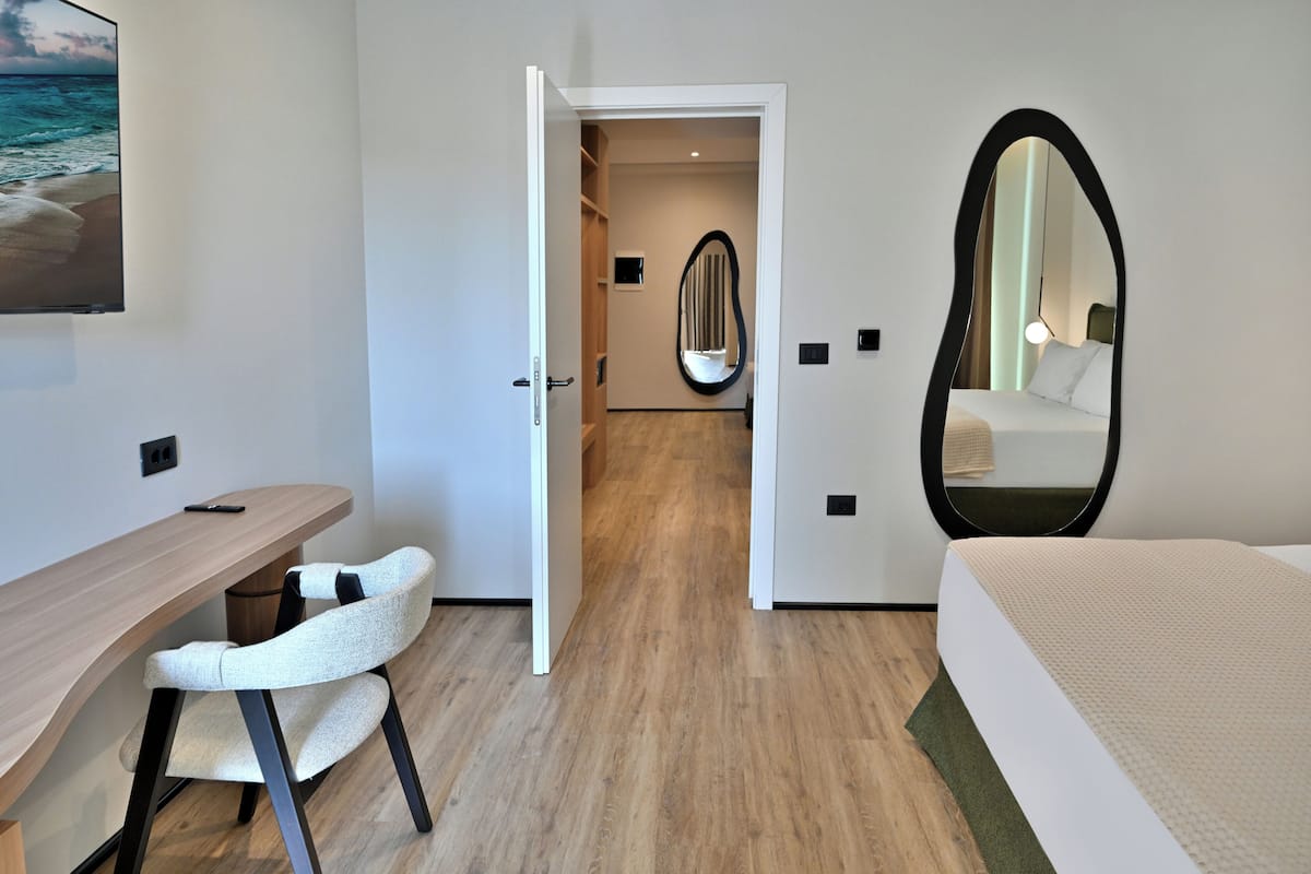 A hotel room with a wooden desk, a white chair, a bed with a green rug, and a large mirror.