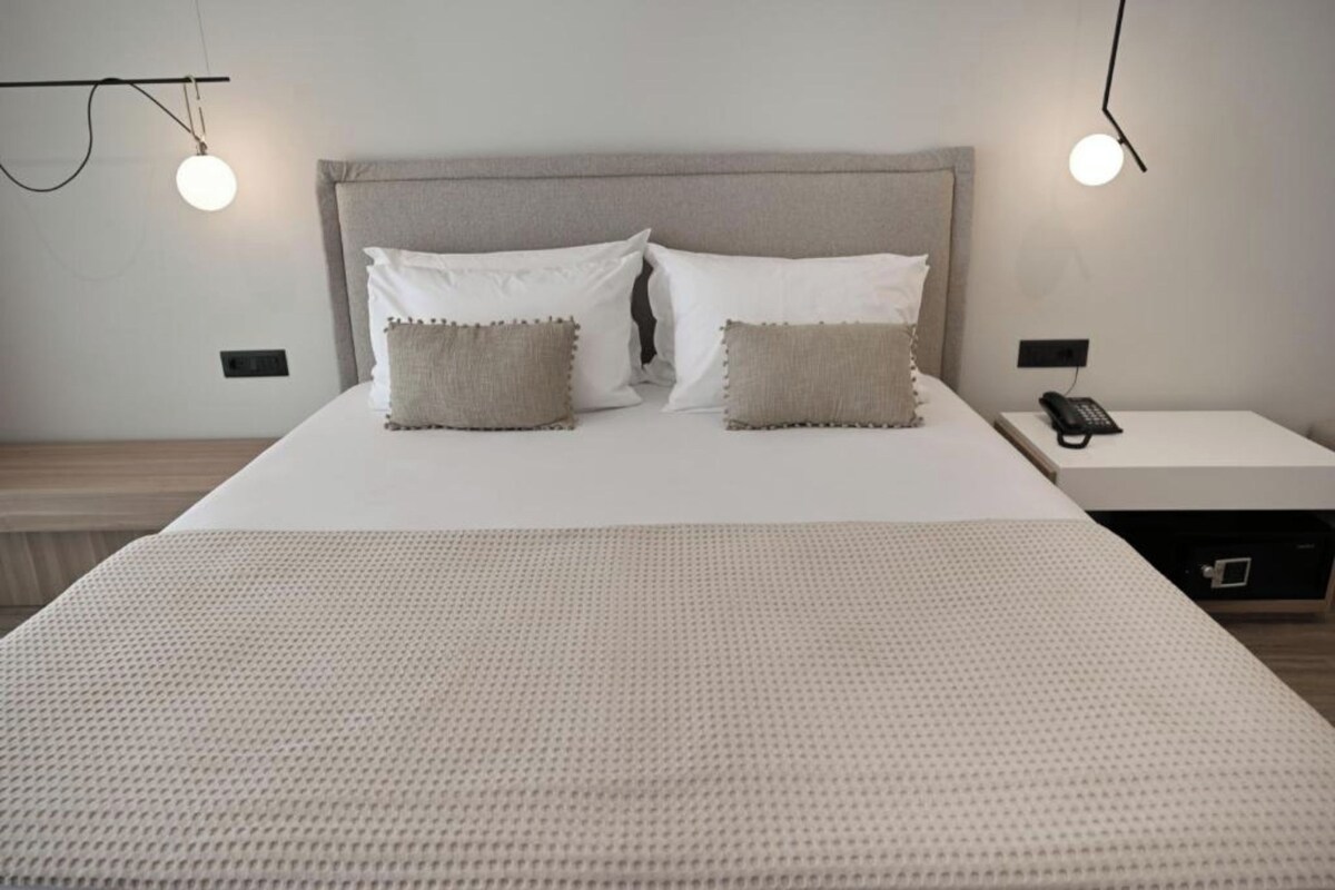 A neatly made bed with a grey headboard, two white pillows, and two beige pillows. There is a bedside table with a black telephone and a small drawer.