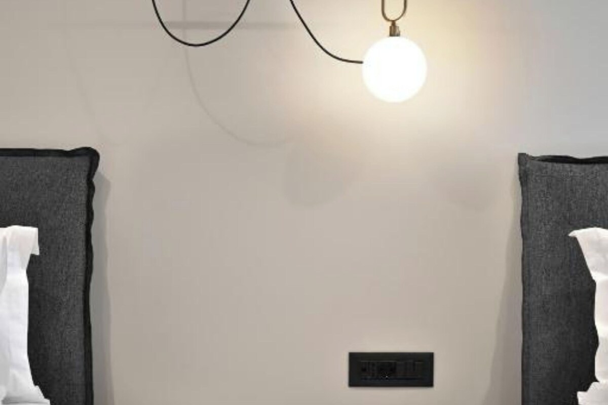 A modern bedroom with a white bedside table, a hanging light fixture, and a power outlet on the wall.