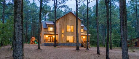 A wooden cabin with a large front porch, surrounded by tall trees.