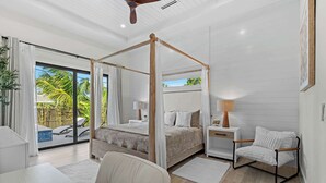 A bedroom with a four-poster bed, a sitting area with a chair, a desk, and a view of a pool and palm trees through large windows. - Brand New Gulf Coast Luxury Getaway (Naples)