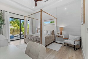 A bedroom with a four-poster bed, a sitting area with a chair, a desk, and a view of a pool and palm trees through large windows.