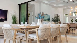 A dining area with a large wooden table surrounded by wicker chairs, white cushions, and a white vase with blue flowers. - Brand New Gulf Coast Luxury Getaway (Naples)