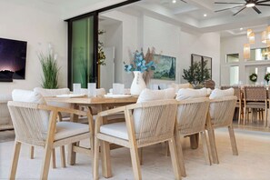 A dining area with a large wooden table surrounded by wicker chairs, white cushions, and a white vase with blue flowers. - Brand New Gulf Coast Luxury Getaway (Naples)
