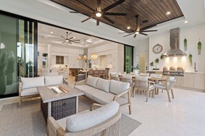 A spacious living area with a large sofa, a coffee table, and wicker chairs. There is a kitchen in the background with modern appliances and a dining area with wooden tables and chairs.