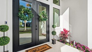 A modern house entrance with a black door, two wreaths, a welcome mat, and a small garden with shrubs and flowers. - Brand New Gulf Coast Luxury Getaway (Naples)