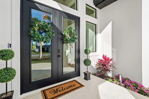 Une entrée moderne avec une porte noire, deux couronnes décoratives, un paillasson et un petit jardin agrémenté d’arbustes et de fleurs.