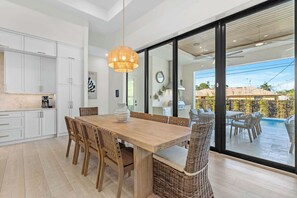 A modern kitchen with white cabinets, a wooden dining table, wicker chairs, and a large window overlooking a pool. - Brand New Gulf Coast Luxury Getaway (Naples)