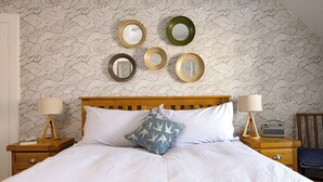 Een net opgemaakt bed met witte lakens, een houten hoofdbord en een blauw kussen met een vogelmotief. Op één nachtkastje staan een lamp en een radio. De muur is bekleed met decoratief golvend behang.